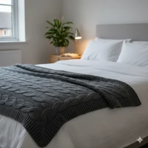 A dark grey cable knit throw folded at the foot of a bed with white linen in a minimalist bedroom.