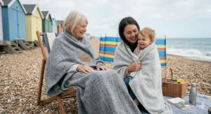 An elderly lady and a young child wrapped in travel blankets on a pebble beach with colourful British beach huts in the background.