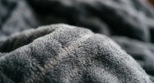 A 4K photorealistic macro photograph showing the intricate soft texture and fibres of the grey fleece fabric used in a Beurer heated blanket, with subtle internal heating wires just visible, illuminated by natural light.