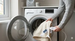 A detailed photorealistic 4K image showing hands placing a Silentnight electric blanket into a washing machine drum. The detached control unit is resting clearly in the foreground, demonstrating that the blanket is machine washable. Natural light illuminates the soft fabrics, matching the clean aesthetic of the main scene.