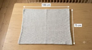 A technical flat lay diagram showing the spread-out cot blanket on an oak floor with dimensions 100 cm by 75 cm clearly labelled.