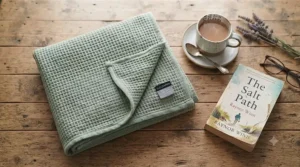 A flat lay image of a breathable cotton-blend lightweight blanket under £20 alongside a cup of tea and a paperback book.
