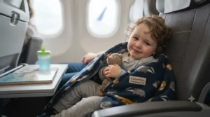 Happy toddler using a cozy travel nap blanket while relaxing in an aeroplane seat during a family holiday flight.
